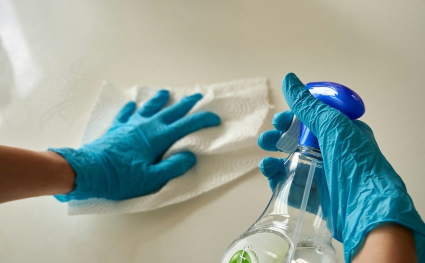 Benefits of Hiring a Professional House Cleaner. Hands wearing blue gloves cleaning a flat surface with a paper towel and spray bottle. 